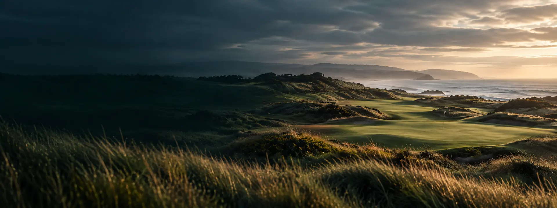 Links dunes before sunrise under heavy cloud, wind-shaped marram grass in the foreground and a fairway falling away towards the sea.